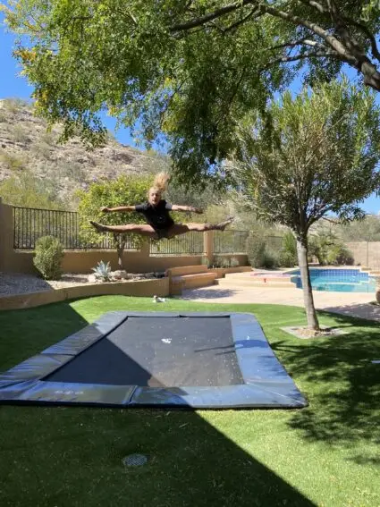 A man joyfully jumps on a trampoline in a sunny backyard, surrounded by green grass and trees.