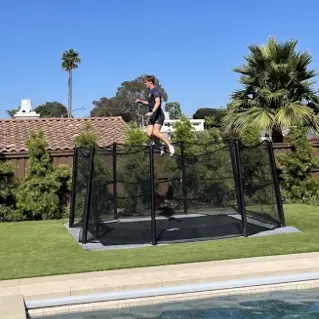 A man joyfully jumps on a trampoline in a sunny backyard, surrounded by grass and trees.